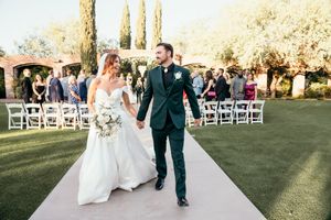 Bride and groom in dark green suit walk hand-in-hand down outdoor wedding aisle after ceremony.