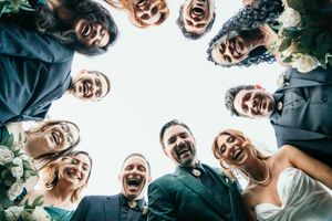 Wedding party laughing and looking down at camera in a fun circular group photo outdoors.