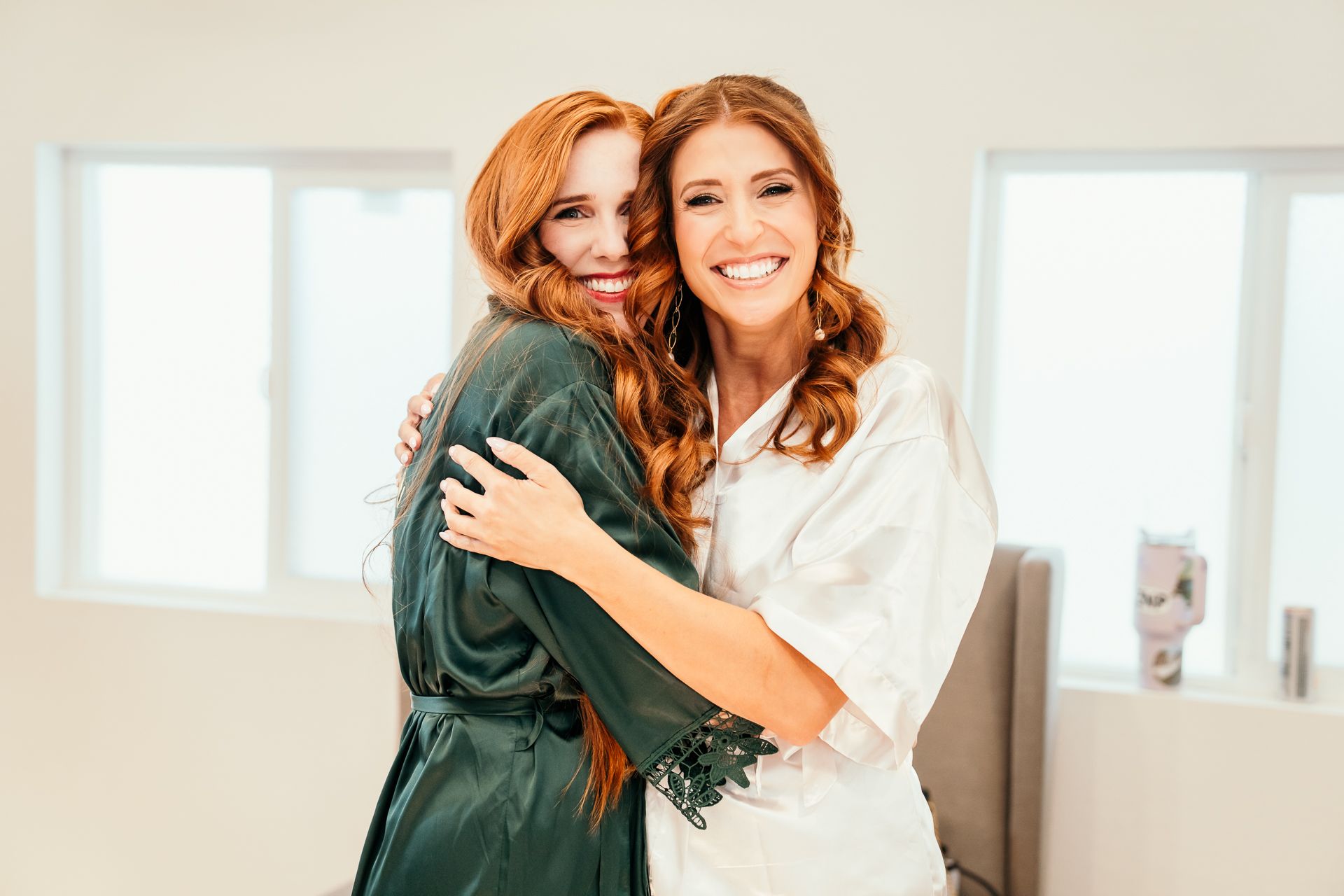 Two women in robes embracing and smiling, one in green satin and one in white lace, posing together indoors.
