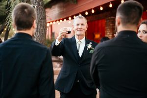 Older man in dark suit raises a glass in a toast at an outdoor wedding reception near a brick building.
