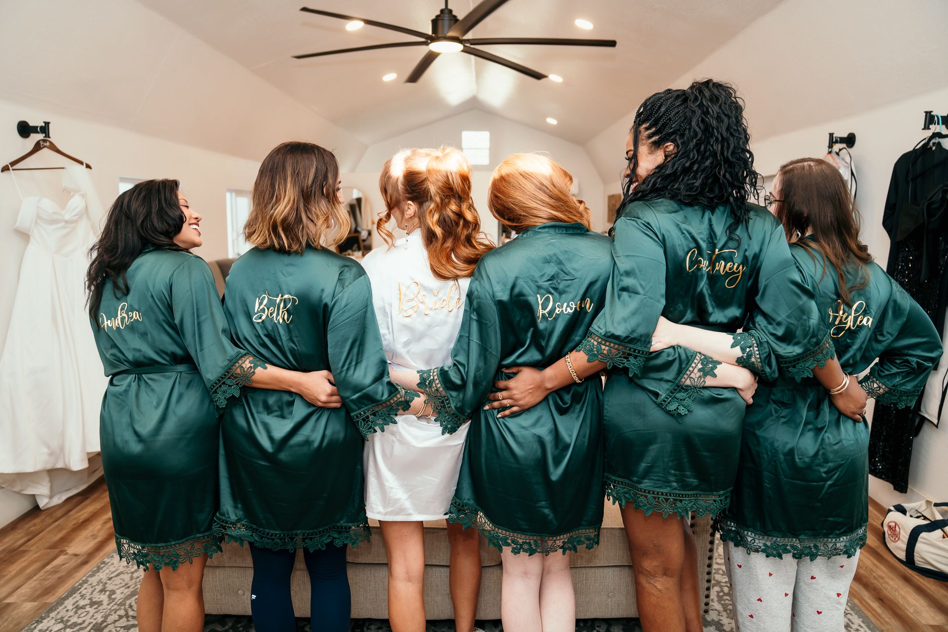 Bride and bridesmaids in personalized emerald green satin robes standing together with backs to camera.