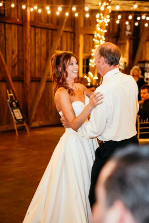 Bride in strapless white gown shares father-daughter dance in warmly lit barn wedding reception.