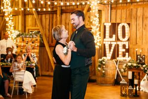 Couple shares a romantic mother-son dance in a rustic barn wedding venue with string lights and a LOVE sign.