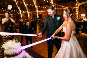 Bride and groom cut wedding cake with glowing lightsabers in a rustic barn reception with string lights.