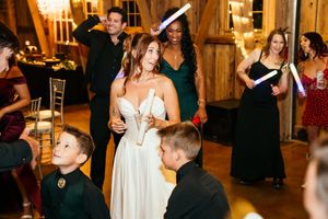 Bride in white gown celebrates with guests holding glow sticks on wedding reception dance floor.