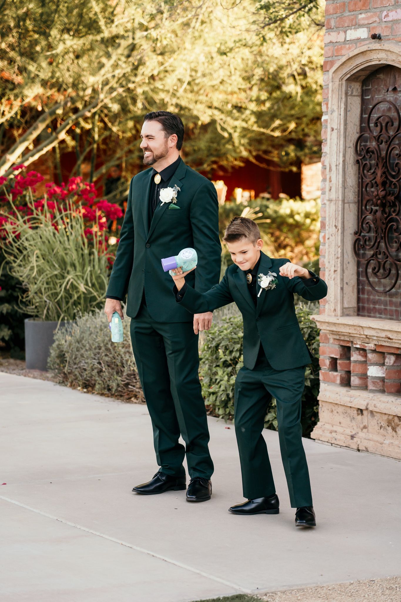 Man and boy in matching dark green suits with boutonnieres walk together at an outdoor wedding ceremony.