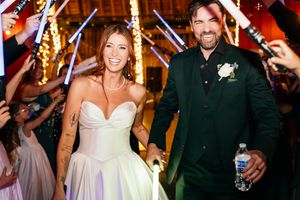 Bride and groom exit wedding reception through a lightsaber tunnel, smiling and holding hands in elegant attire.