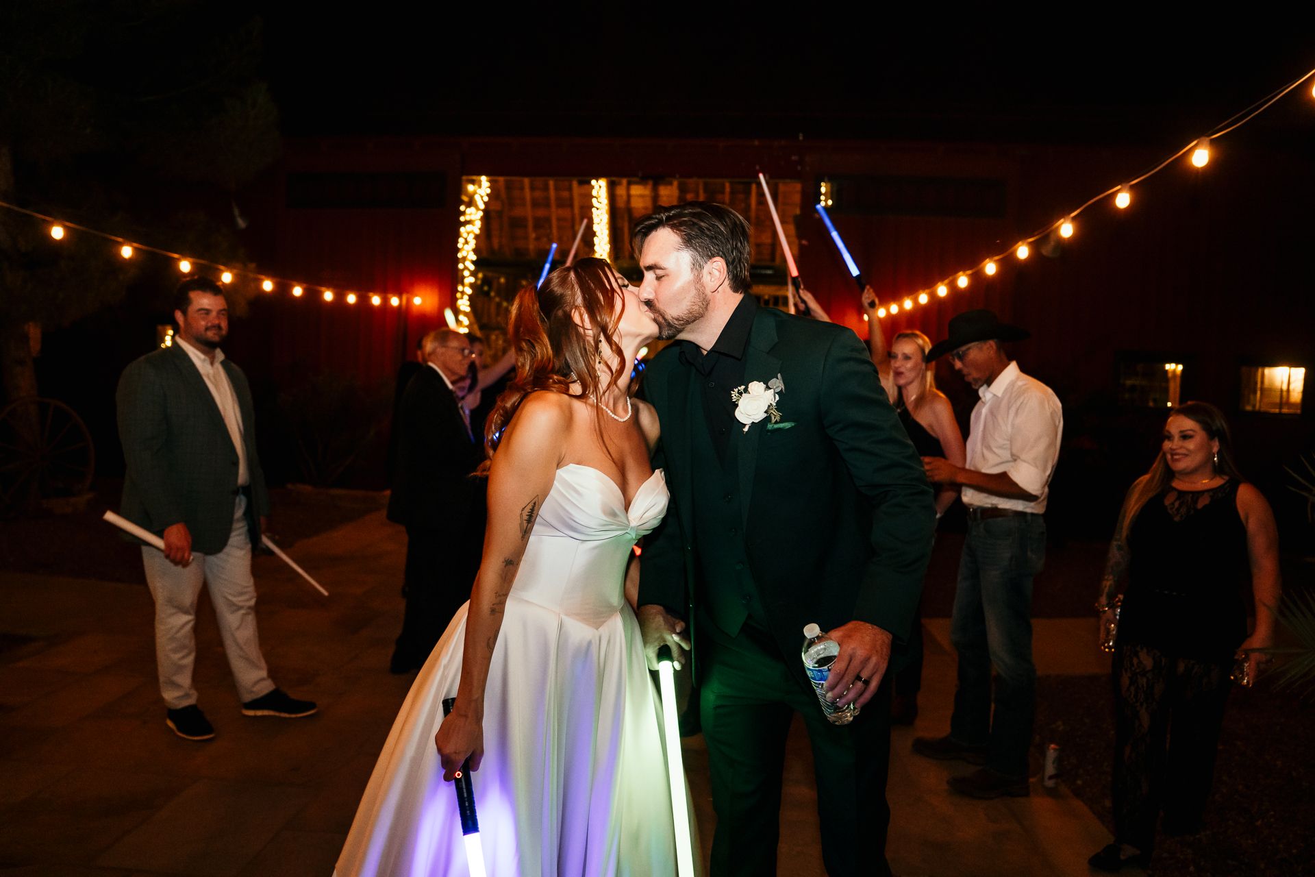 Bride and groom share a kiss surrounded by guests holding lightsabers at outdoor night reception.