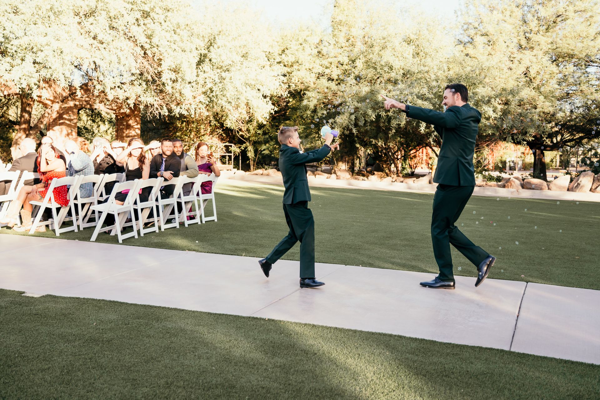 Two groomsmen in dark suits high-five on a grass aisle at an outdoor wedding ceremony with white chairs.
