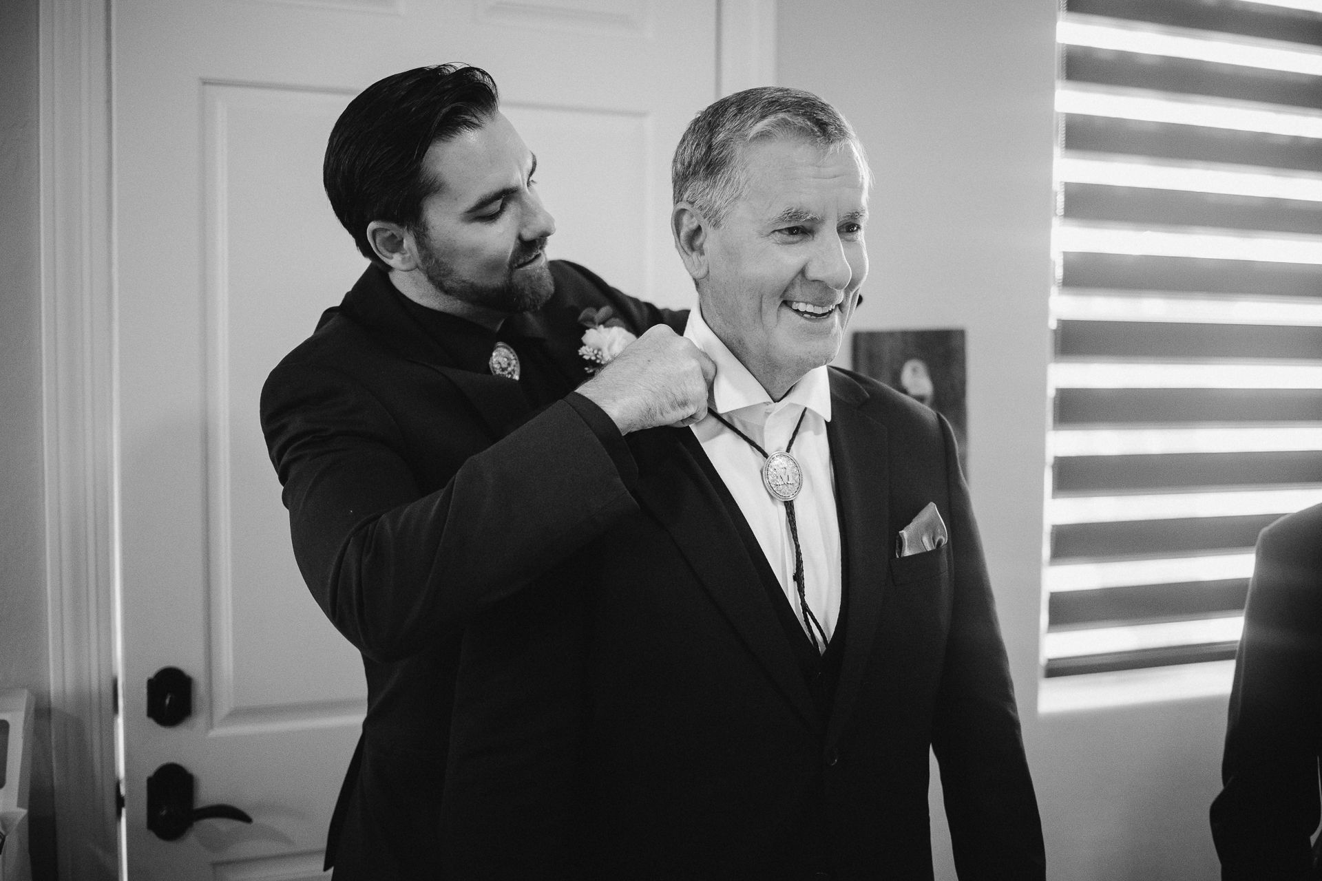 A man helps adjust another mans suit jacket in a black and white wedding preparation photo.