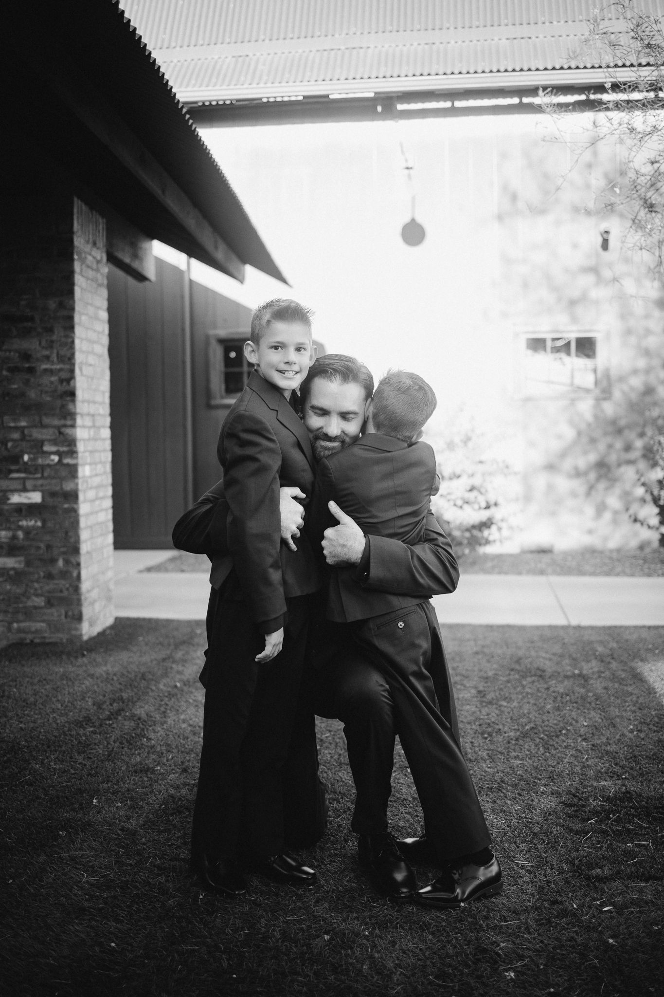 Black and white photo of groom hugging two young boys in suits outside wedding venue.