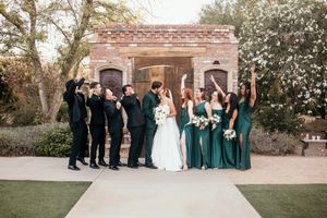 Wedding party in emerald green and black attire celebrate outside brick venue with blooming trees.