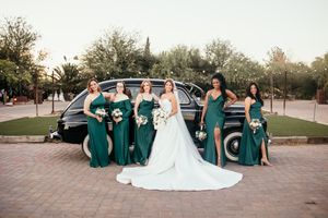 Bride and four bridesmaids in emerald green gowns pose in front of a vintage black car.