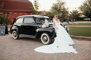 Bride in white gown poses beside a classic black vintage car on brick pavement at an outdoor wedding venue.