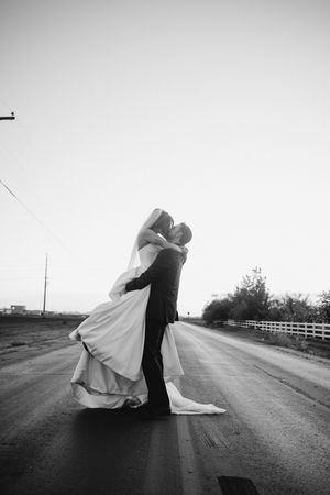 Black and white photo of groom lifting bride for a kiss on an empty road on their wedding day.
