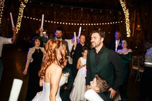 Bride and groom share a joyful dance with guests holding glow sticks at a rustic barn wedding reception.