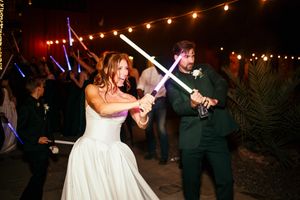 Bride and groom battle with glowing lightsabers at outdoor Star Wars themed wedding reception.