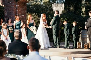 Outdoor wedding ceremony with bride and groom exchanging vows, bridesmaids in emerald green, and flower girls under a chandelier.