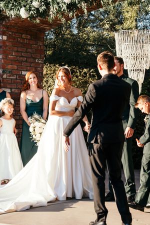 Smiling bride in off-shoulder white gown faces groom at outdoor wedding altar with bridesmaids in emerald green.