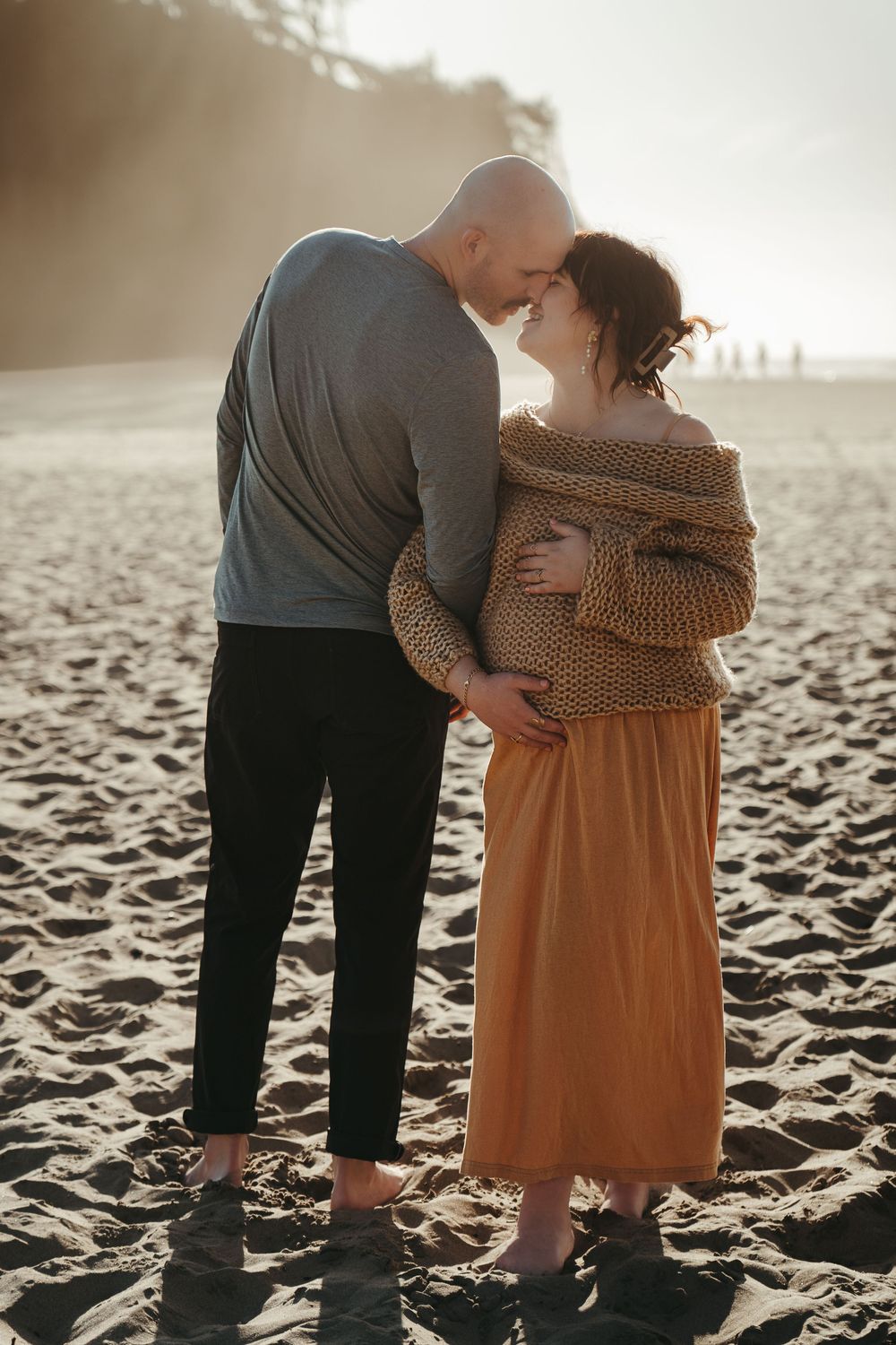 Expecting couple sharing a tender kiss on a misty beach at golden hour.