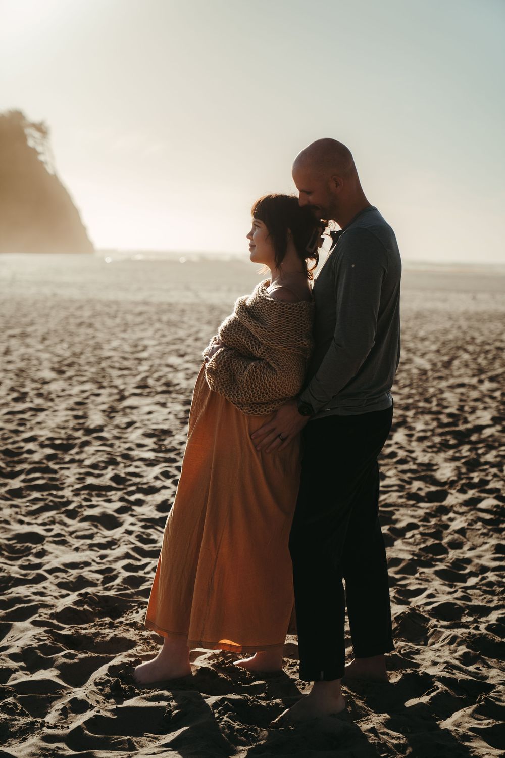 Pregnant woman and partner embracing on sandy beach in warm sunset light.