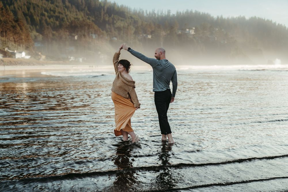 Couple dancing joyfully in shallow ocean water on a misty Pacific Northwest beach.