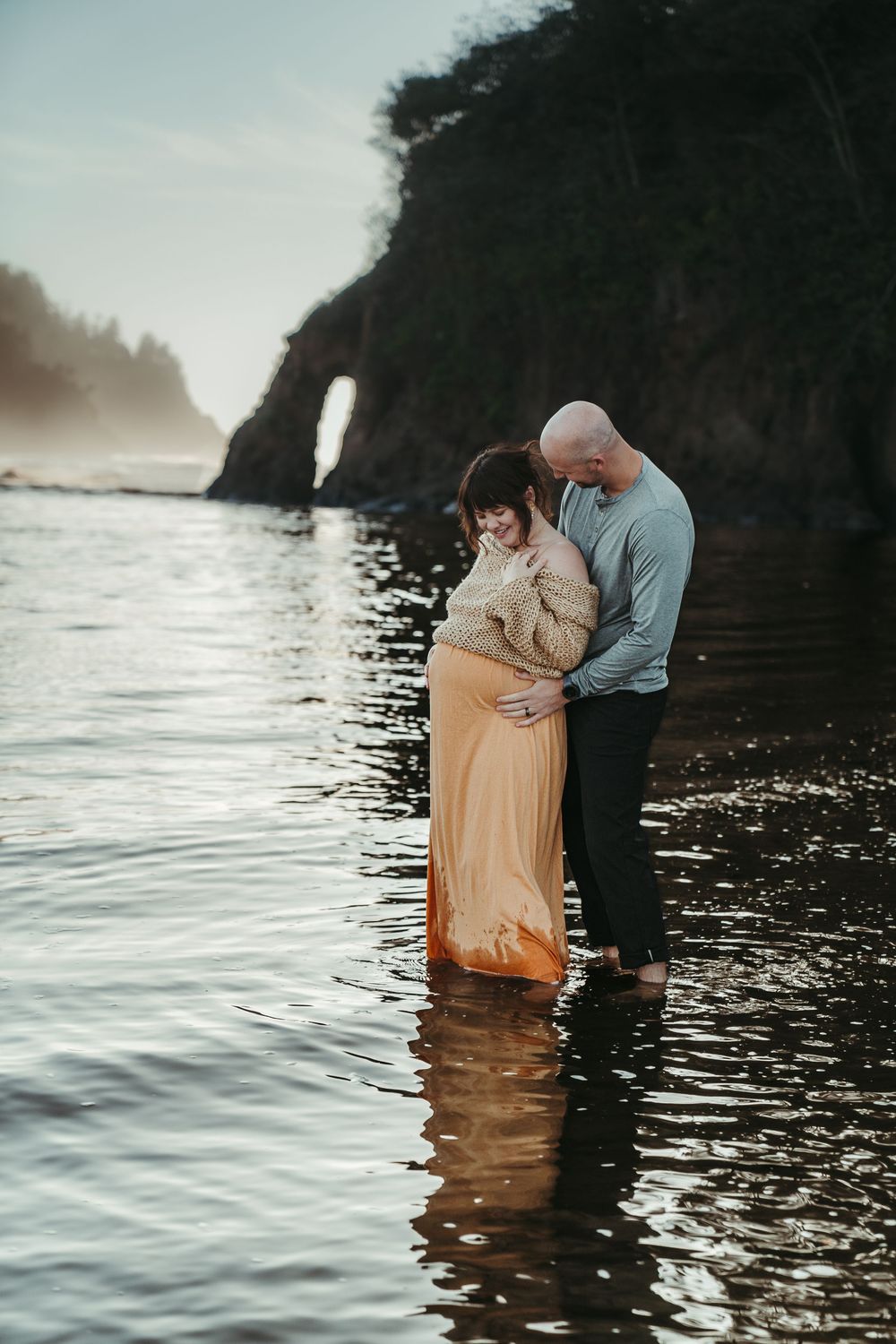 Couple standing in ocean water admiring pregnant belly at scenic coastal location.