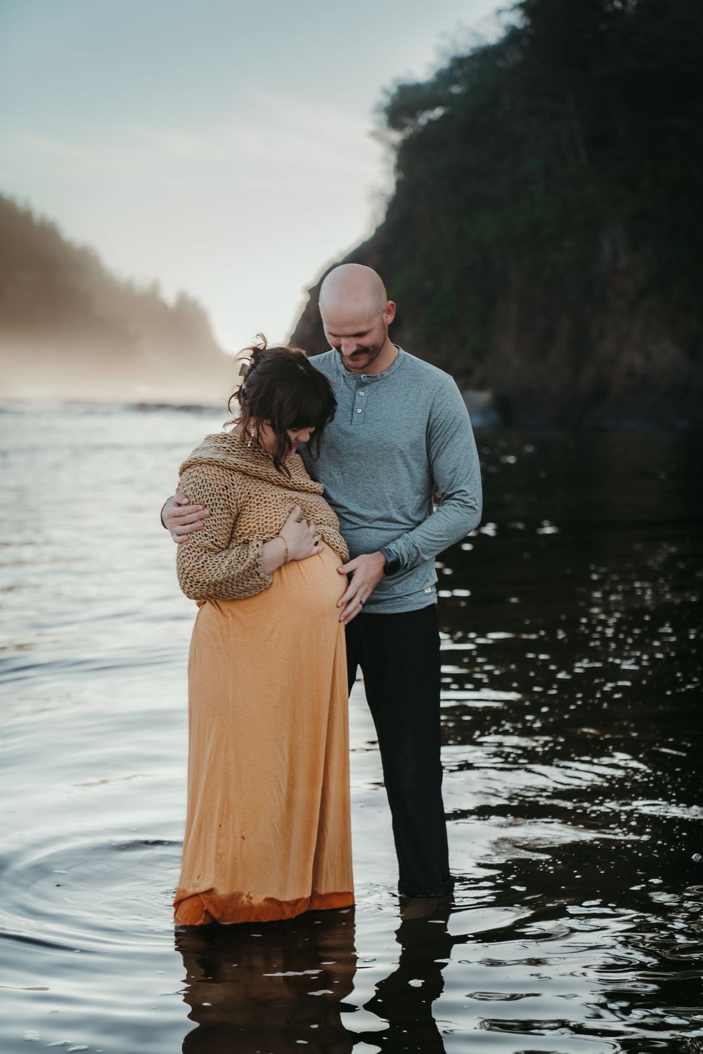 Bald man tenderly embracing pregnant partner standing in calm ocean water at dusk.