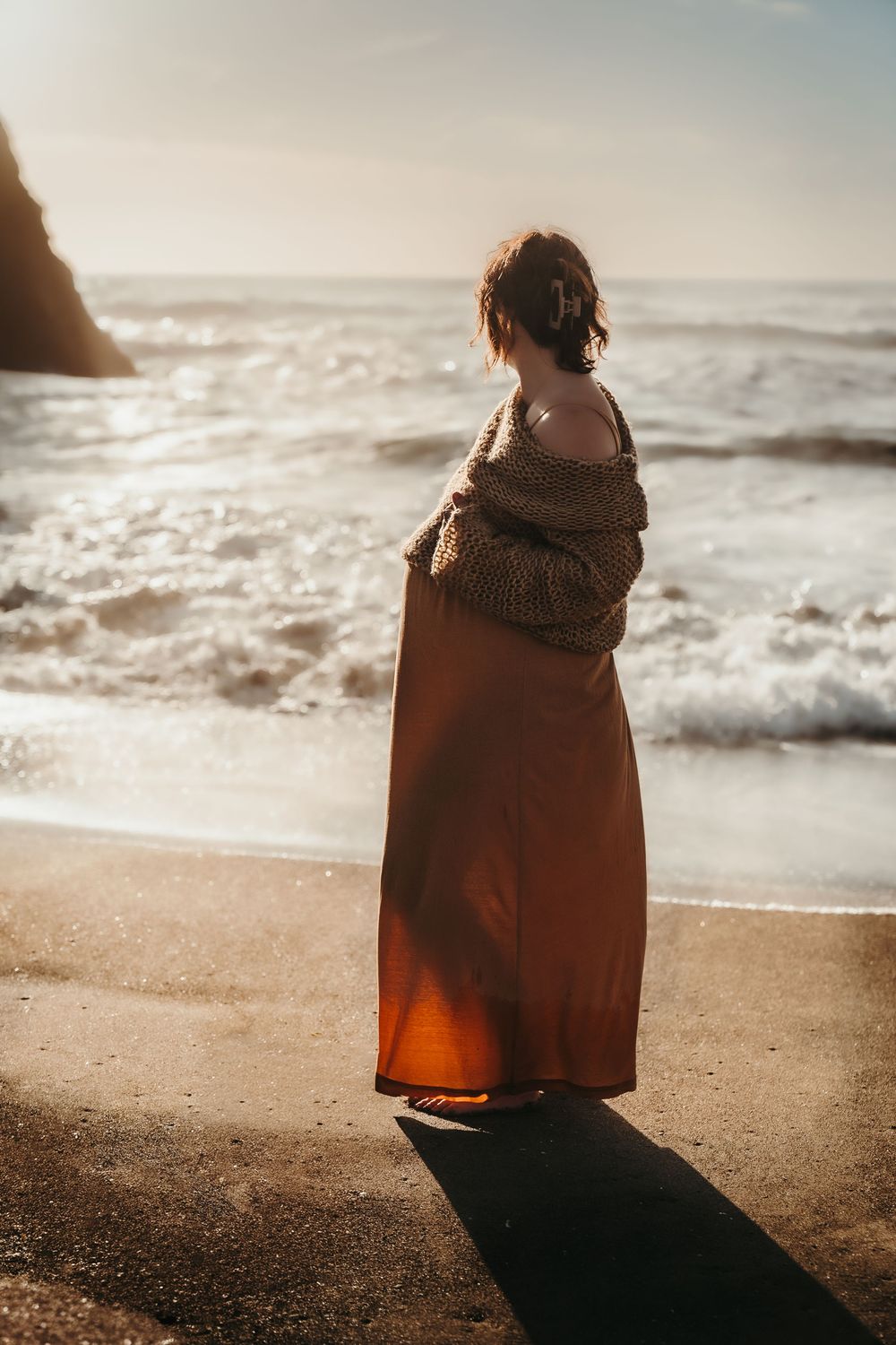 Pregnant woman in orange maxi skirt gazing at ocean waves at golden hour.