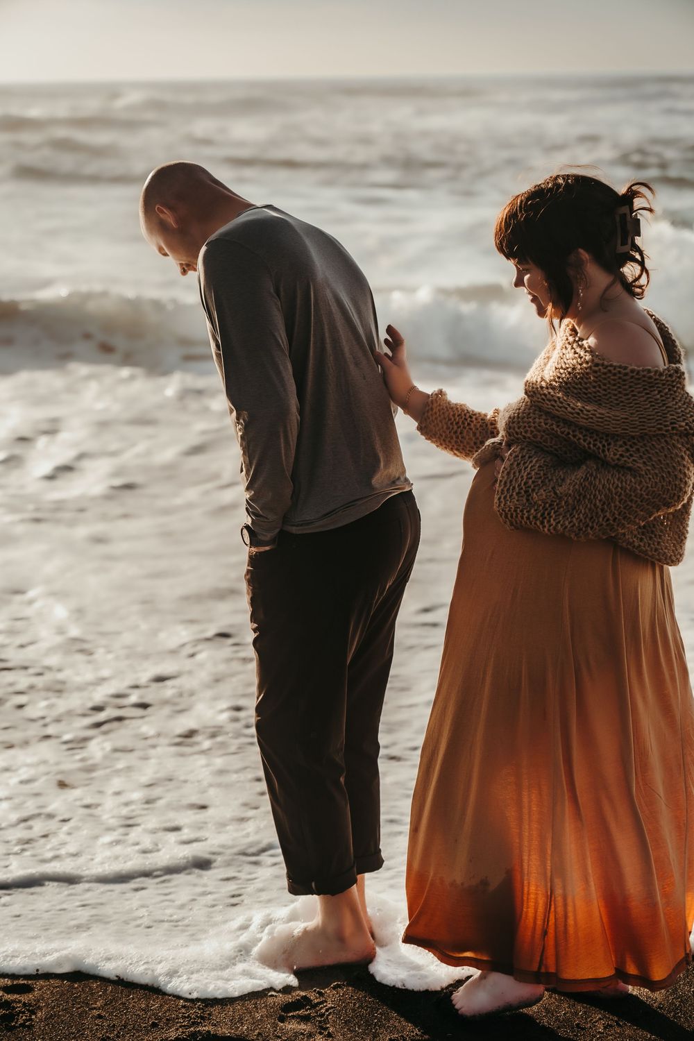 Pregnant woman holding partners hand near crashing waves on a sunlit beach.