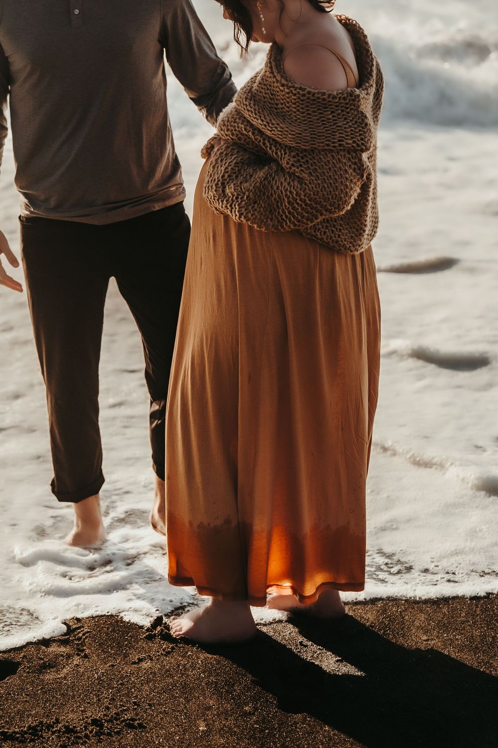 Pregnant woman in rust orange dress and knit shawl standing barefoot in ocean waves with partner.