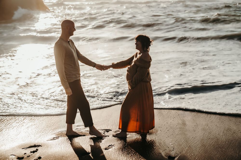Couple holding hands on sunlit beach at golden hour, pregnant woman in orange dress and floral crown.