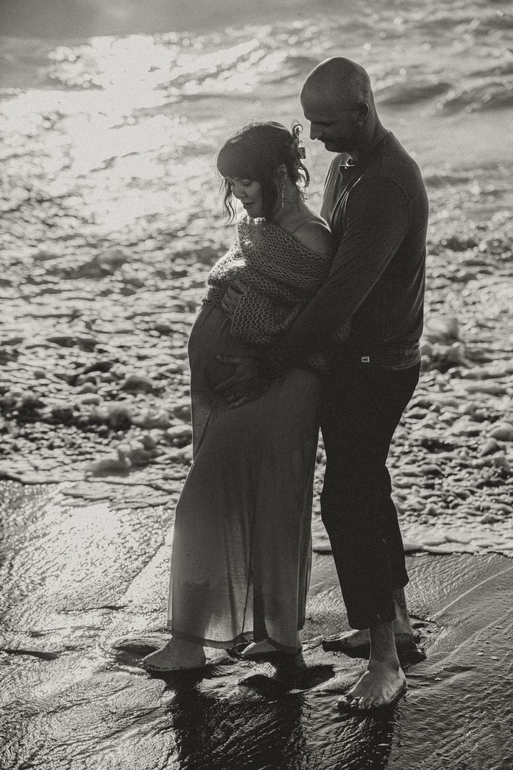 Black and white photo of couple embracing on beach, man tenderly holding pregnant partners belly.