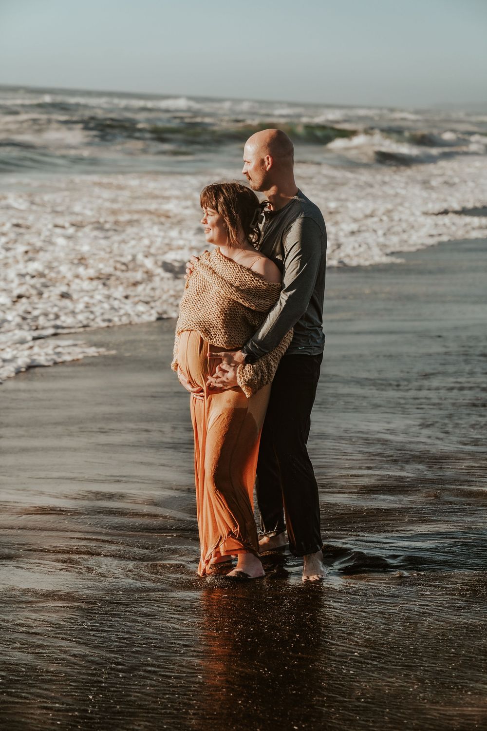 Couple embracing on wet sandy beach with ocean waves, pregnant woman in rust dress and knit wrap.