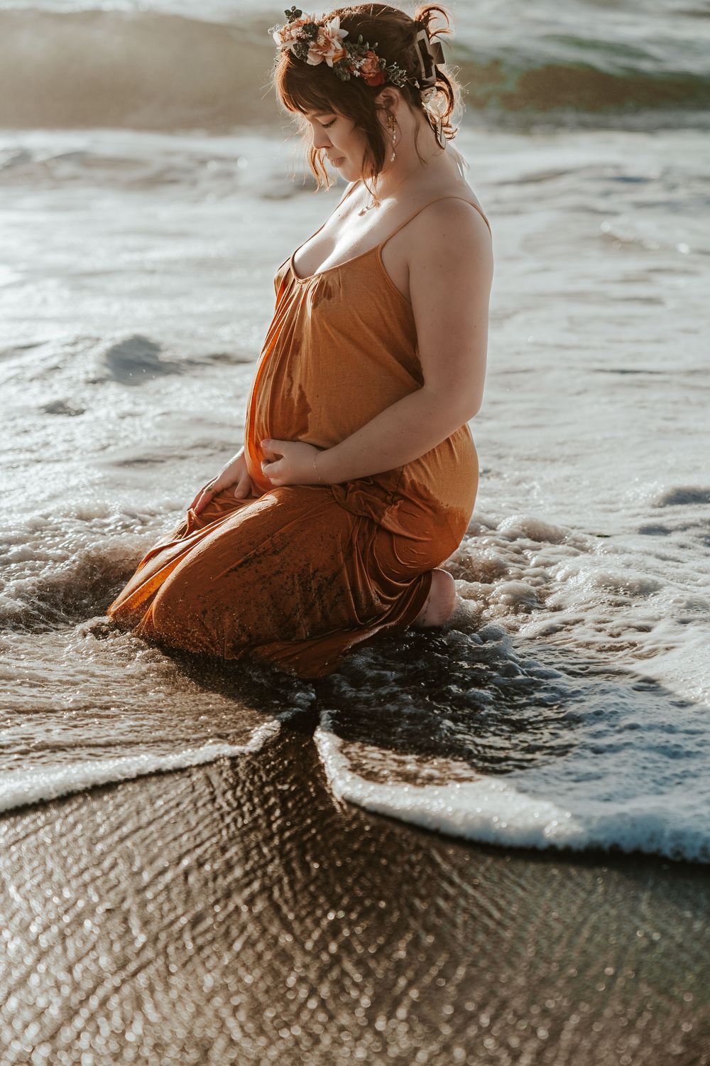 Pregnant woman in rust orange dress and floral crown kneeling peacefully in shallow ocean waves.