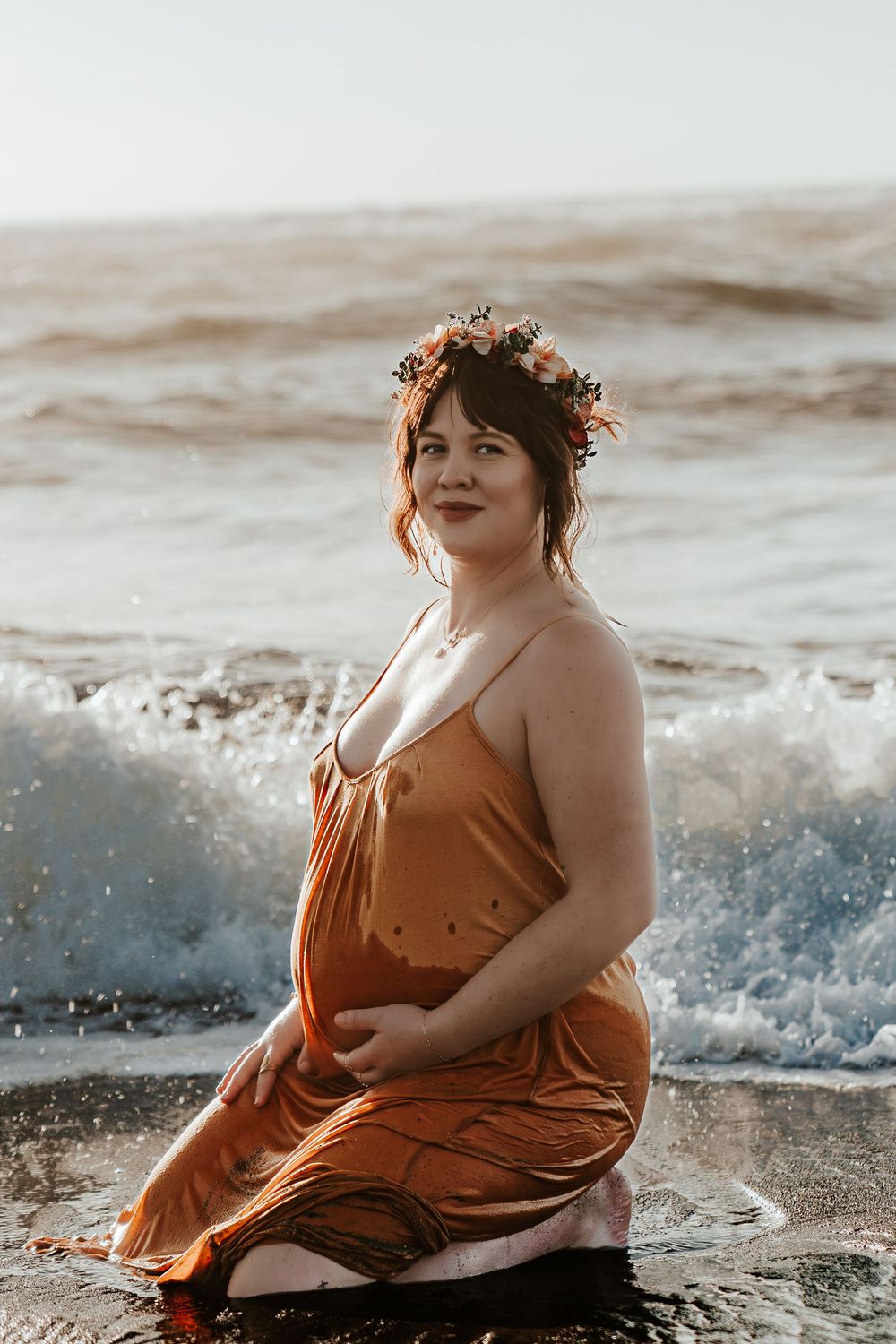 Smiling pregnant woman in orange dress and flower crown sitting in ocean surf on sandy beach.