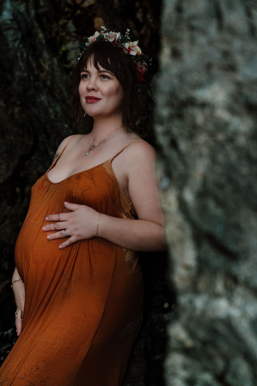 Pregnant woman in rust orange dress and floral crown posing beside rocky surface in natural light.