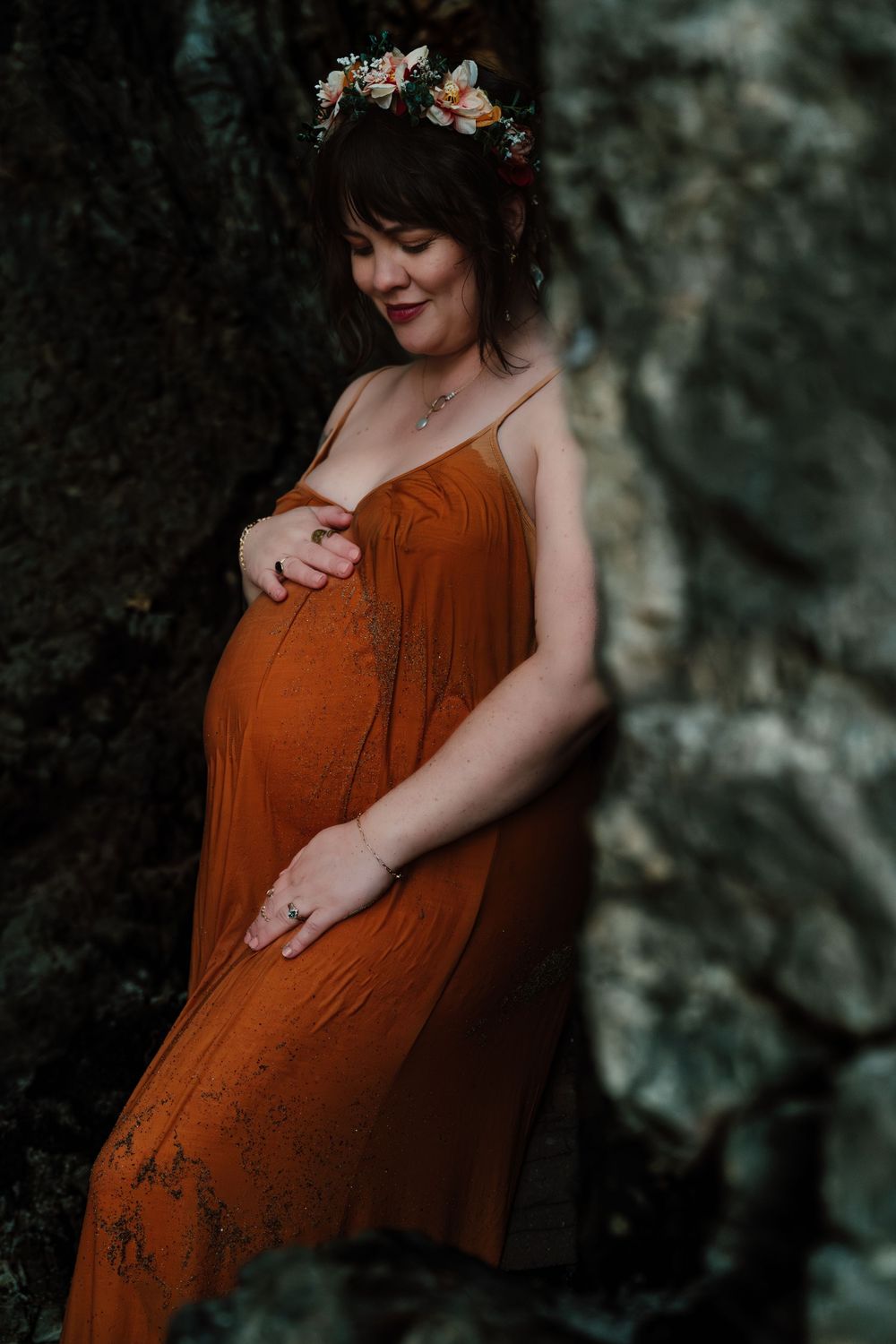 Pregnant woman in burnt orange dress and flower crown cradling belly beside mossy rock wall.