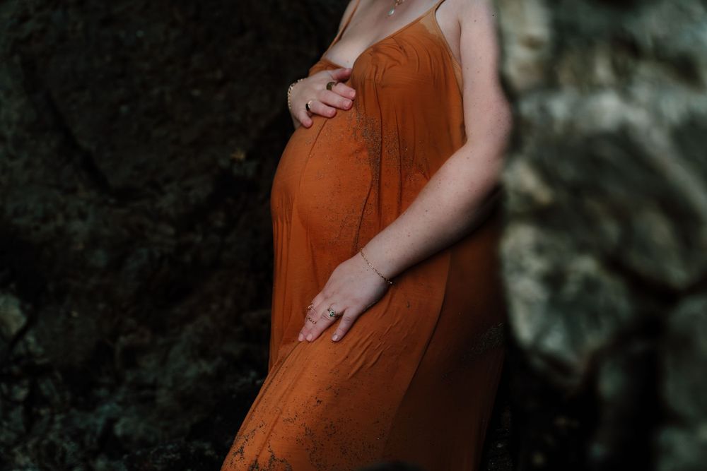 Close-up of pregnant womans baby bump in rust orange dress with hands gently resting on belly.