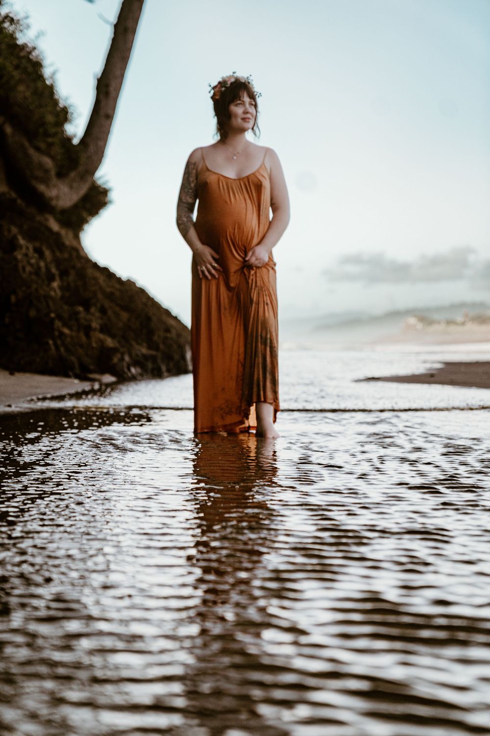 Pregnant woman in rust orange maxi dress wearing floral crown standing in shallow beach water at sunset.