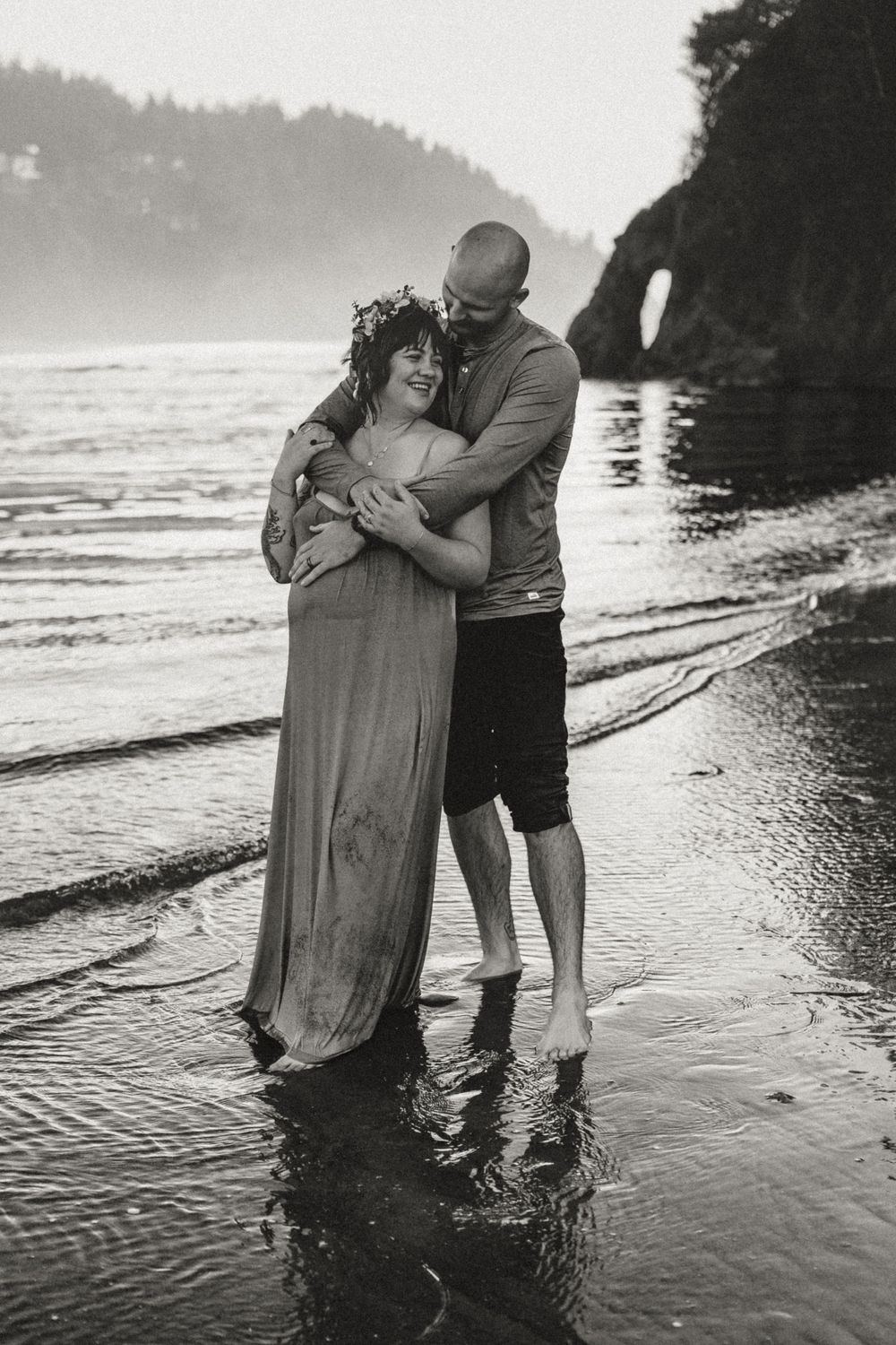 Black and white photo of a couple embracing on a misty beach, woman in flowing dress wearing a floral crown.