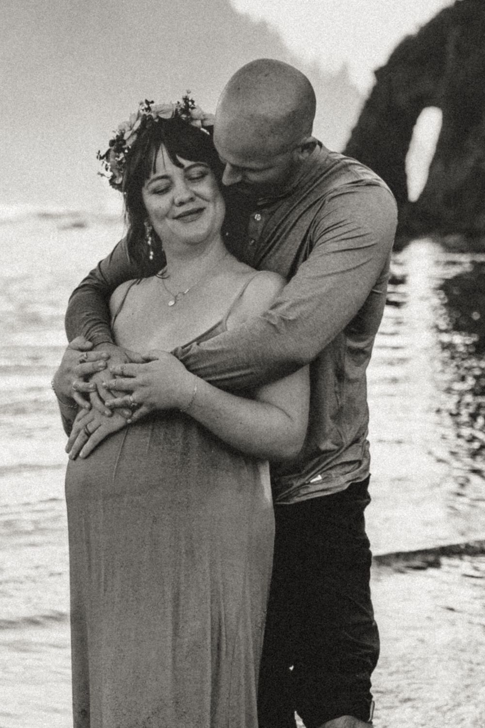 Black and white maternity photo of couple embracing on rocky beach, woman wearing floral crown.
