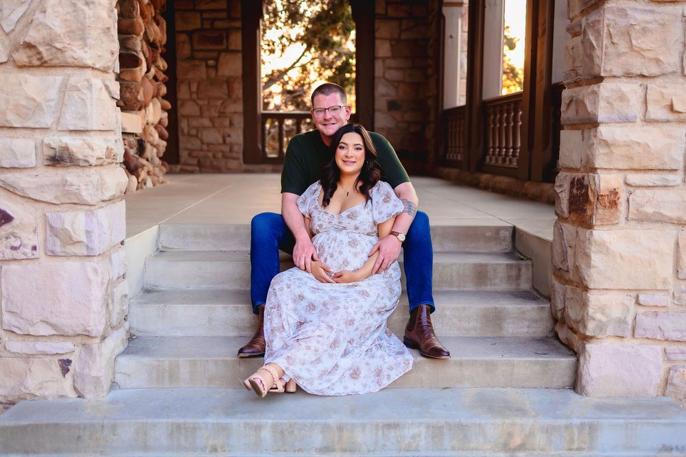 A pregnant woman in a floral dress sitting on stone steps with a man.  They are possing for photos in a stone archway corridor.