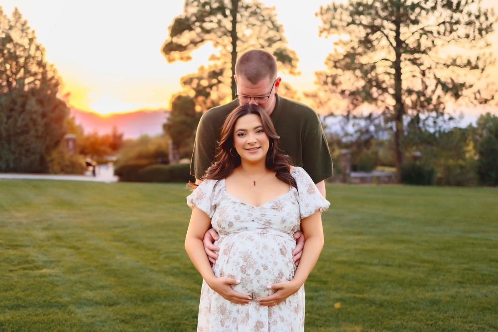 A couple embraces in a grassy field during golden hour sunset as warm light illuminates the landscape.  Woman pregnant in floral dress