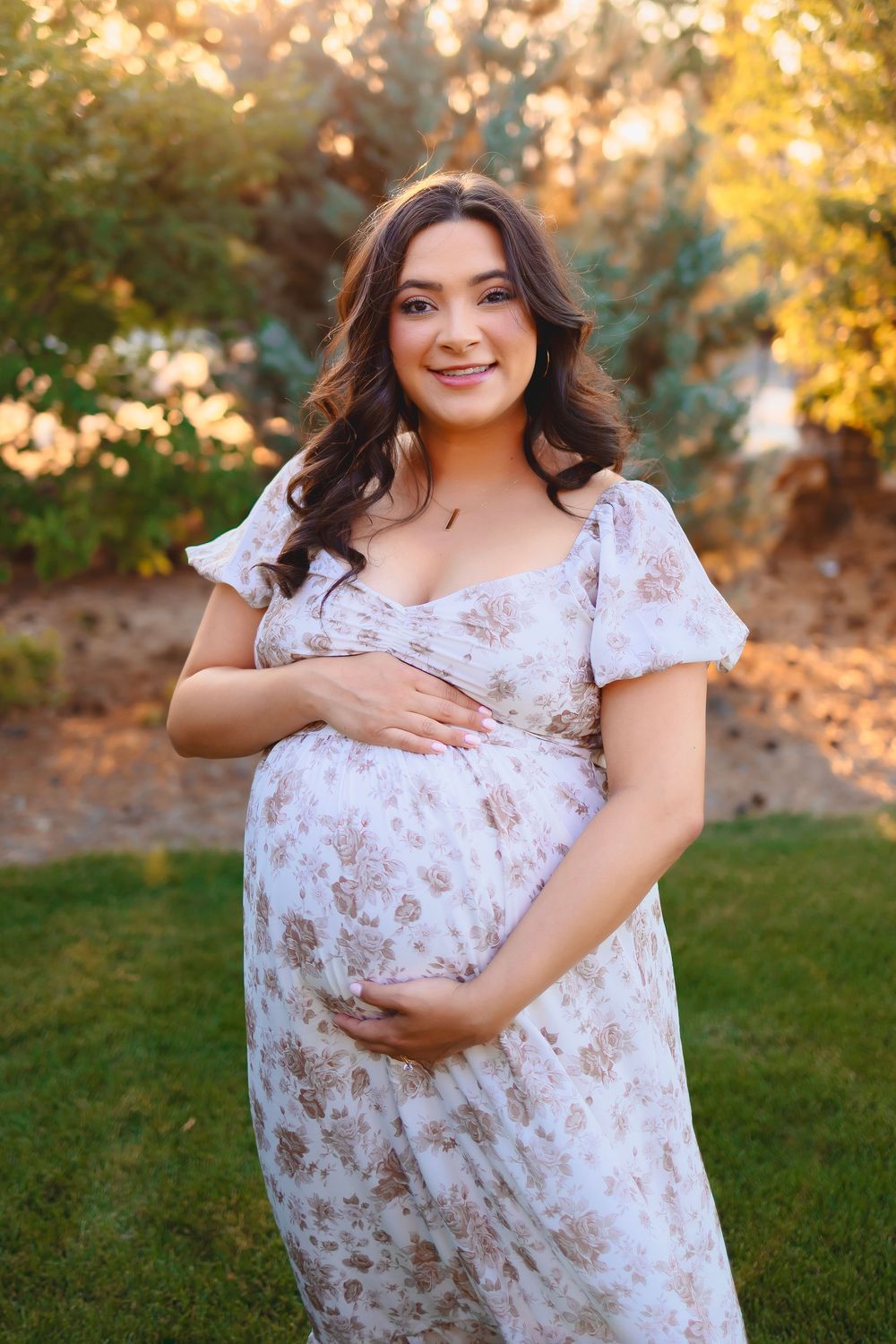 A maternity photo session in a park with golden autumn lighting shows a woman in a white floral dress.