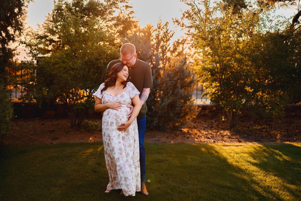 A couple embraces in a golden sunset-lit field with trees creating a warm, romantic outdoor setting.  Maternity photoshoot highlands ranch