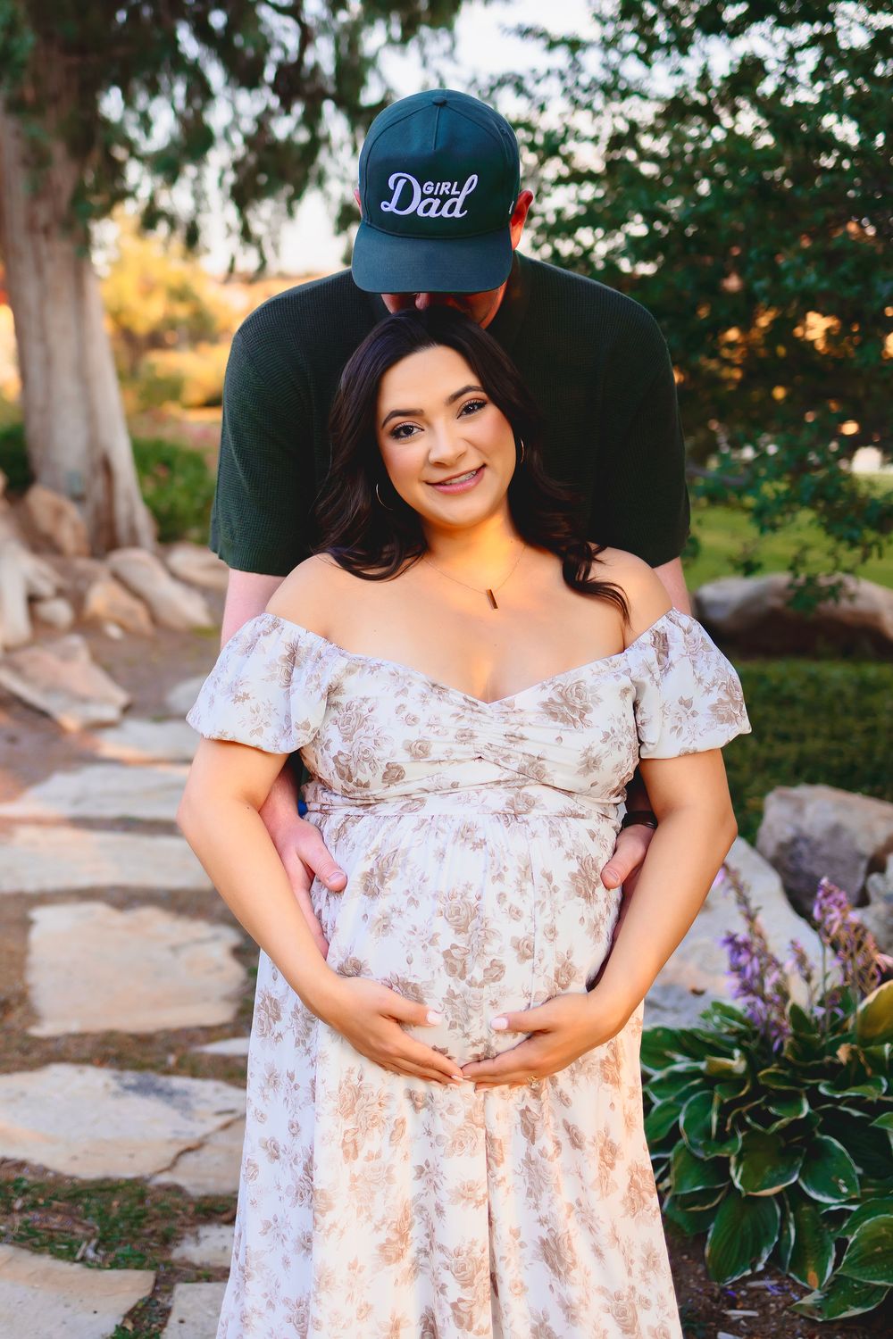 Expectant couple poses in garden setting with woman wearing floral off-shoulder dress and partner in baseball cap saying girl Dad.