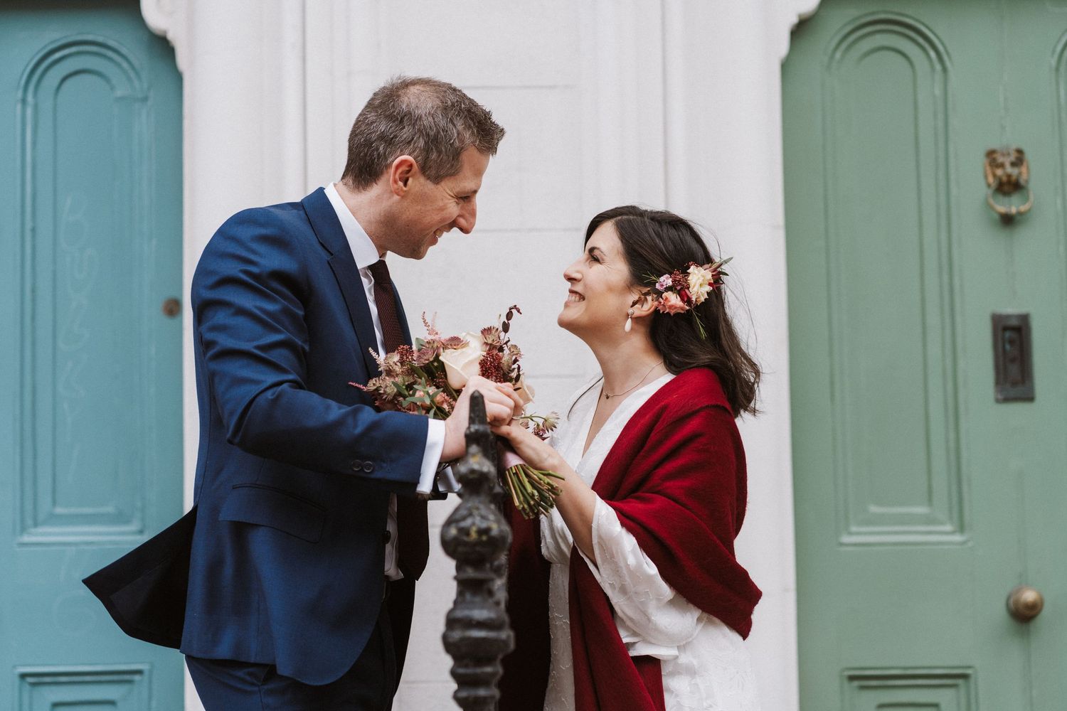 A couple in wedding attire share a laugh while holding drinks in front of turquoise doors.