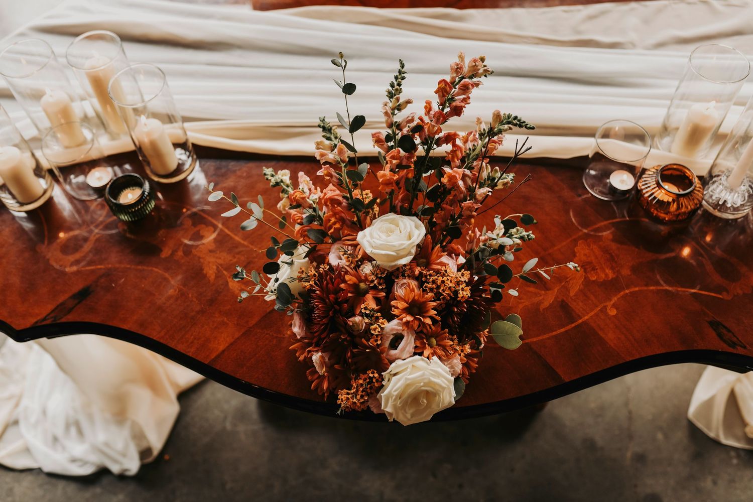 a close up photo of a decorative vintage table draped with an ivory tapestry and covered in tall glass vases with ivory candles