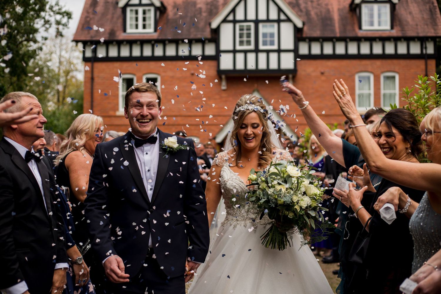 Wedding guests shower confetti on a couple outside a Tudor-style building.